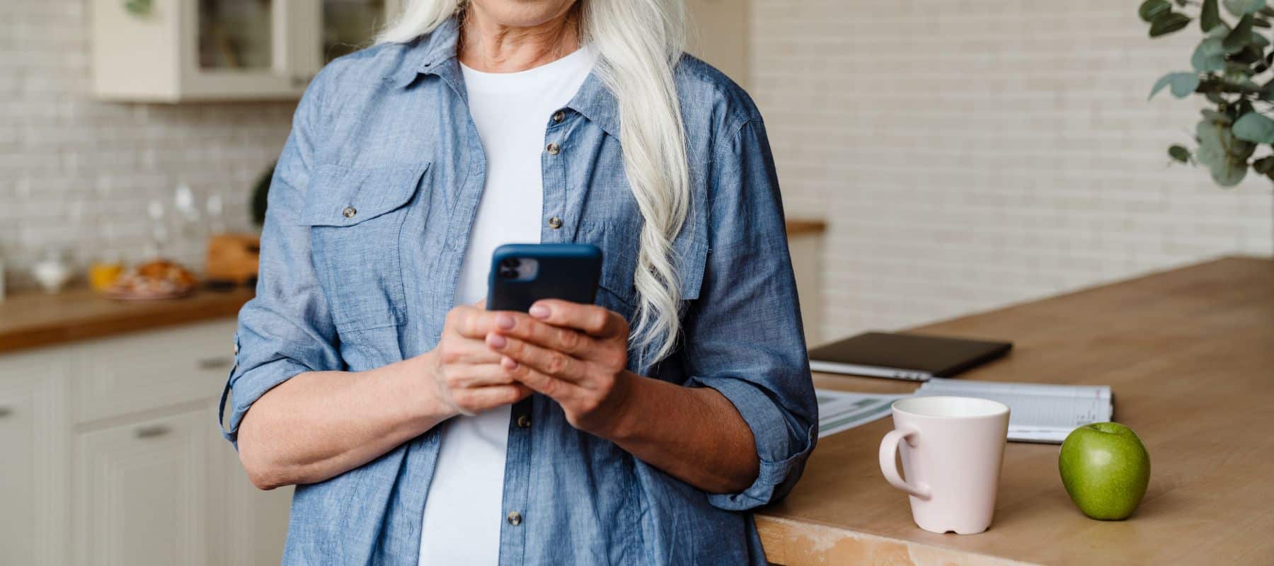 Middle-aged woman with long blonde hair wearing a blue denim shirt, using a smartphone in a modern kitchen setting. A coffee mug and a green apple are visible on the counter beside her, indicating a casual, domestic environment