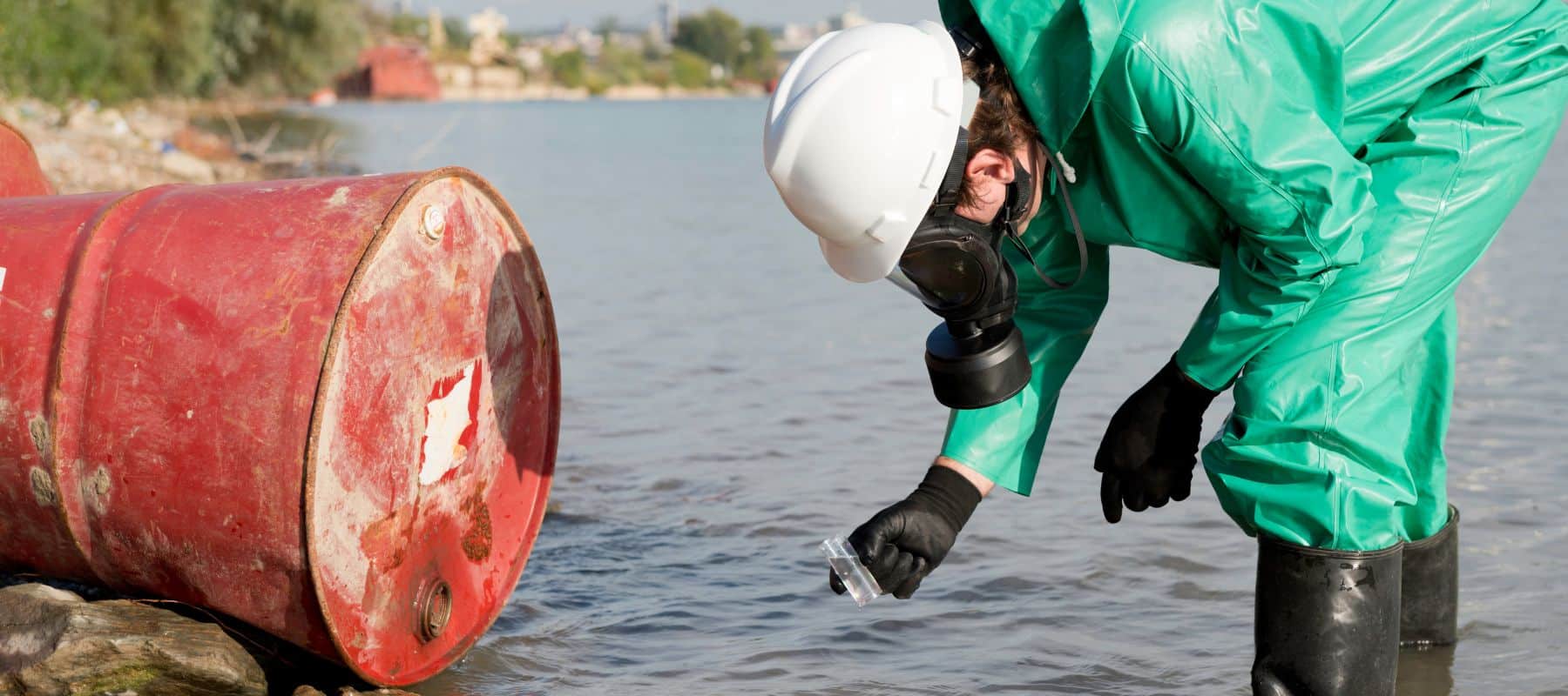 Environmental scientist in a green protective suit and white helmet using a magnifying glass to examine a water sample near a rusty barrel on the shore of a lake, illustrating water quality testing