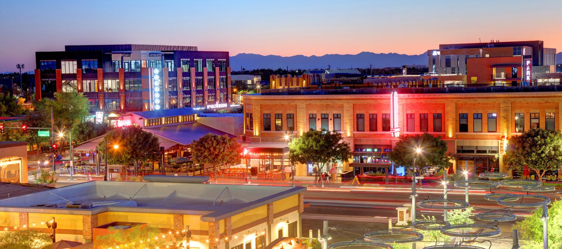 Twilight cityscape showing a vibrant downtown with a mix of modern and historic buildings, illuminated by streetlights and neon signs, against a backdrop of distant mountains under a dusky sky